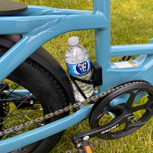 May include: A light blue bicycle frame with a black water bottle holder. A clear plastic water bottle with a blue and white label is in the holder. The bicycle has black tires and a black chain and pedals.