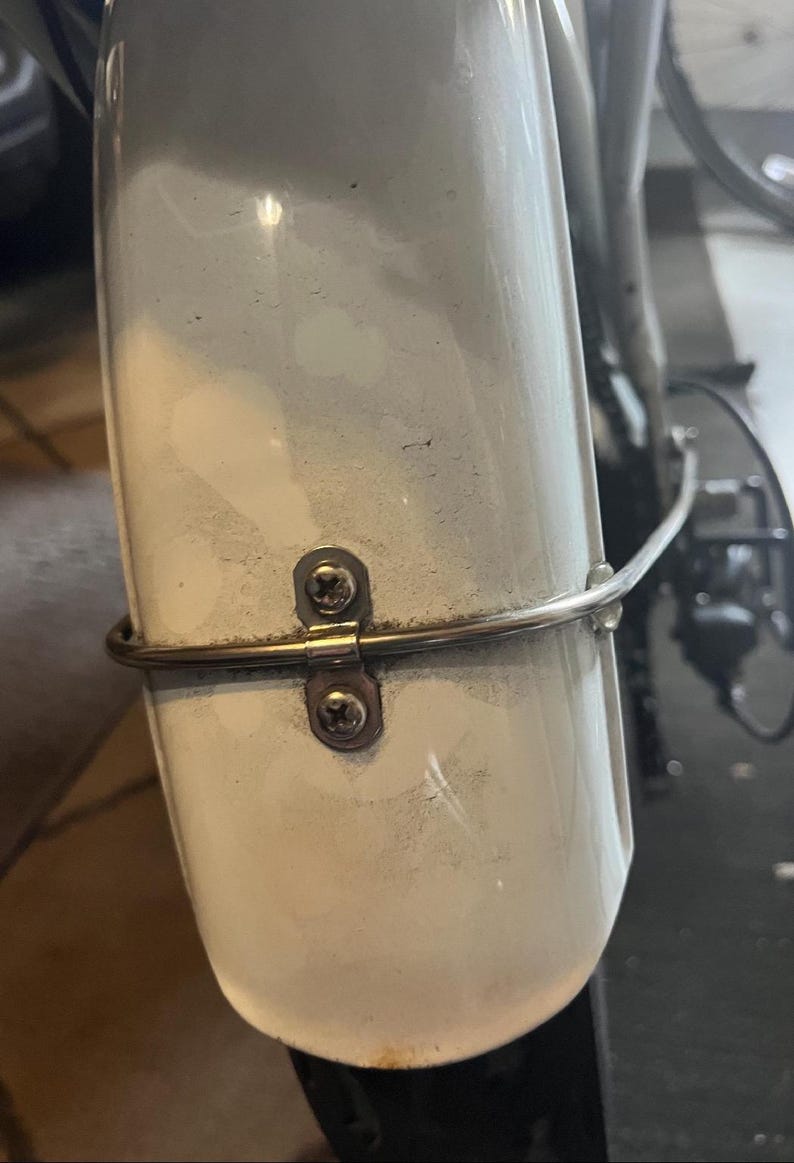 May include: White bicycle fender with a silver metal bracket attached to it with two screws. The bracket has a curved metal bar attached to it.