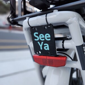 May include: A white bicycle with a black license plate that says "See Ya" in teal blue letters. The bicycle has a red rear reflector.