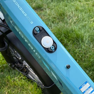 May include: Close-up of a light blue electric bicycle frame with the word "EXPERIENCE" printed on the side. A black component with a white circular device is attached to the frame. The bicycle is on a grassy surface.