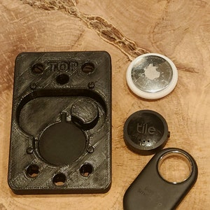 May include: A black 3D printed holder with the word "TOP" printed on it. The holder has several circular cutouts and is designed to hold small round objects. There are three small round objects next to the holder: a white round object with a silver Apple logo, a black round object with the word "tile" printed on it, and a black key-shaped object with the words "Go Find My" and "Smart Tag" printed on it.