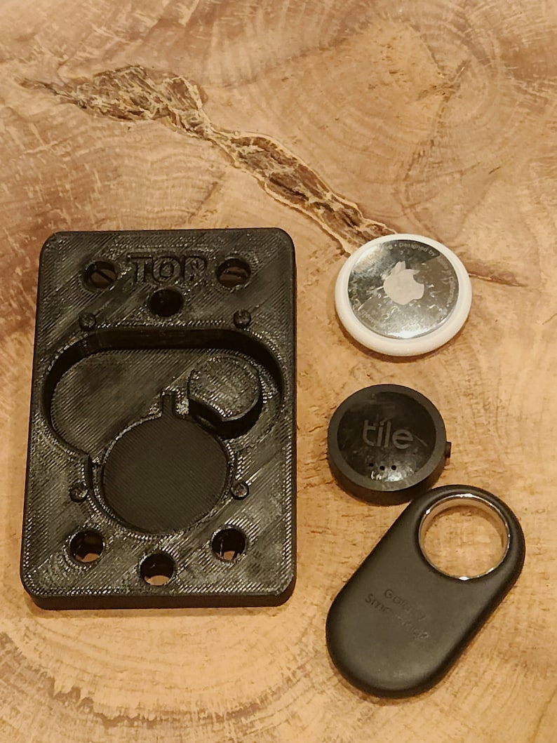 May include: A black 3D printed holder with the word "TOP" printed on it. The holder has several circular cutouts and is designed to hold small round objects. There are three small round objects next to the holder: a white round object with a silver Apple logo, a black round object with the word "tile" printed on it, and a black key-shaped object with the words "Go Find My" and "Smart Tag" printed on it.