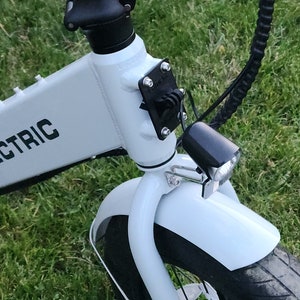 May include: A white electric bicycle with a black front fender, black handlebars, and a black headlight. The bicycle is parked on a green grassy surface.