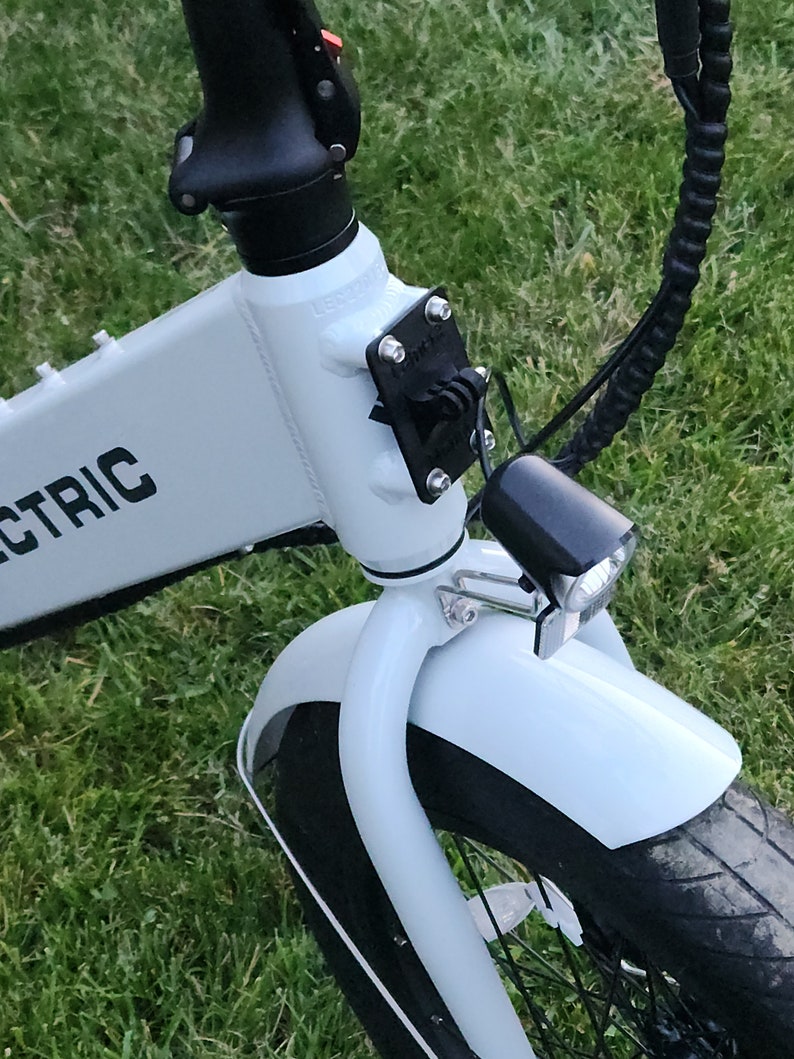 May include: A white electric bicycle with a black front fender, black handlebars, and a black headlight. The bicycle is parked on a green grassy surface.