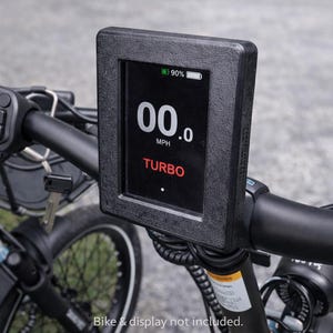 May include: Close-up of an e-bike handlebar with a black display showing "00.0 MPH" and "TURBO" in red. The display indicates 90% battery. The handlebar also features controls and a key.