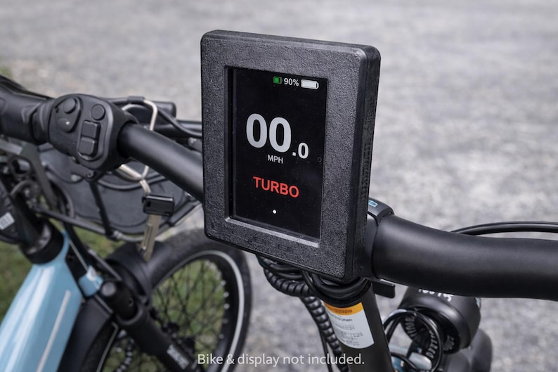May include: Close-up of an e-bike handlebar with a black display showing "00.0 MPH" and "TURBO" in red. The display indicates 90% battery. The handlebar also features controls and a key.