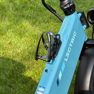 May include: A light blue Lectric e-bike frame with a black water bottle holder. The bike has the word "LECTRIC" printed on the frame. The background is green grass.