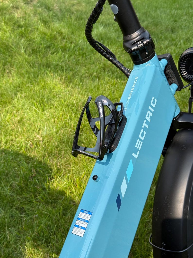 May include: A light blue Lectric e-bike frame with a black water bottle holder. The bike has the word "LECTRIC" printed on the frame. The background is green grass.