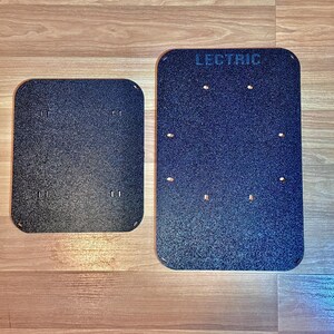 May include: Two black rectangular plates with rounded corners. The larger plate has the word "LECTRIC" printed at the top. Both plates have several small holes and a textured surface. The plates are on a wooden surface.