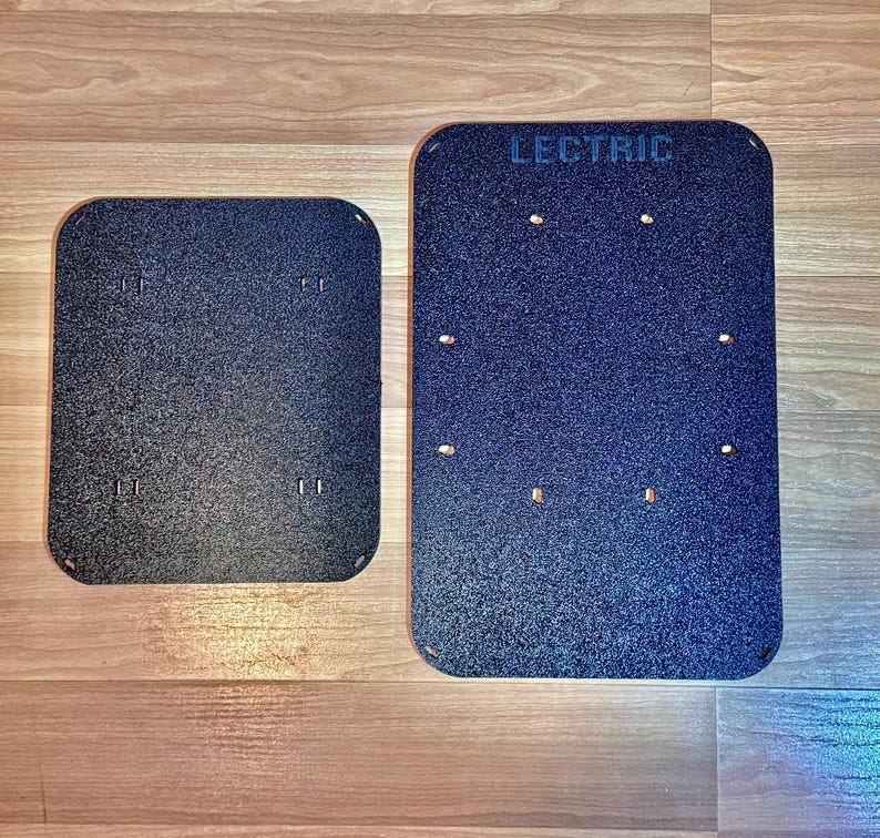 May include: Two black rectangular plates with rounded corners. The larger plate has the word "LECTRIC" printed at the top. Both plates have several small holes and a textured surface. The plates are on a wooden surface.
