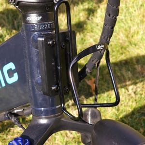 May include: A black bicycle water bottle cage mounted on the frame of a blue electric bicycle. The cage is made of metal and has a simple design. The bicycle has a black handlebar and a black seat.