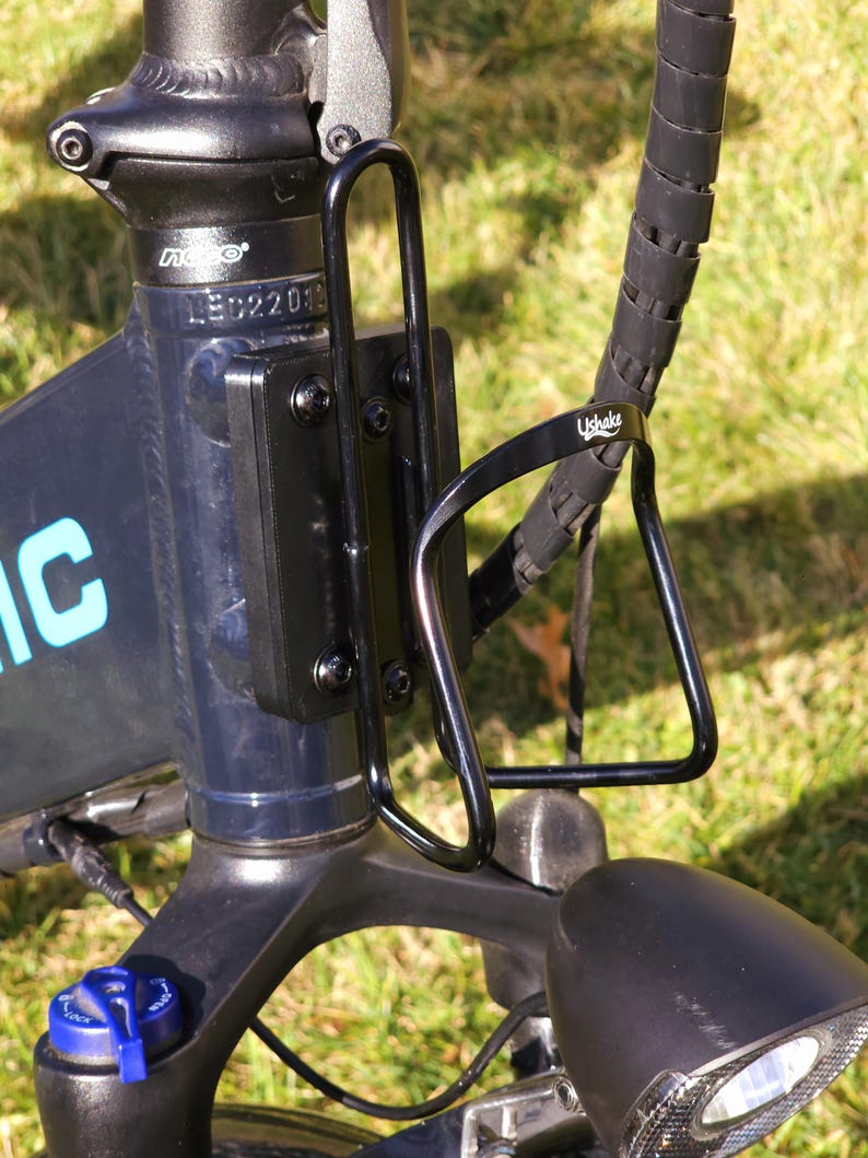 May include: A black bicycle water bottle cage mounted on the frame of a blue electric bicycle. The cage is made of metal and has a simple design. The bicycle has a black handlebar and a black seat.