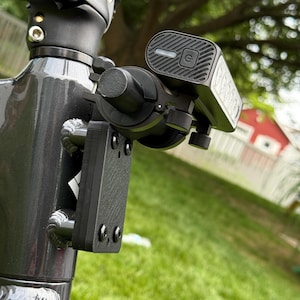 May include: Close-up of a black scooter light and mounting bracket attached to a gray scooter frame. The light has a power button and a charging indicator. The bracket is secured with screws.