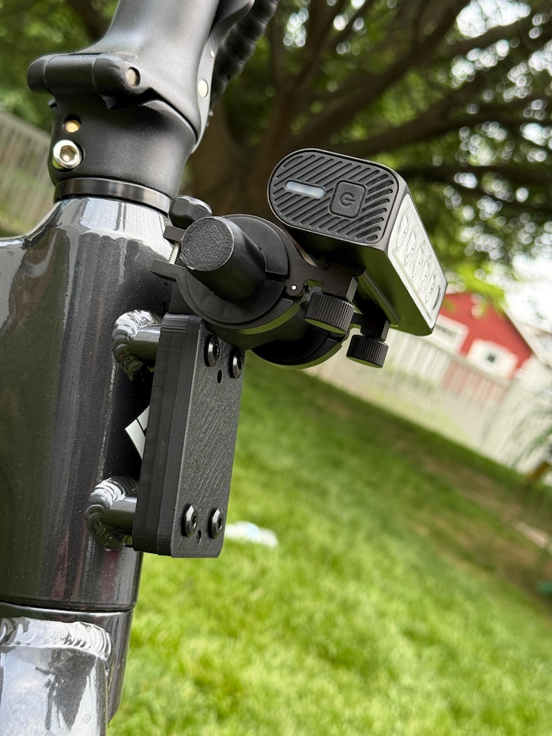 May include: Close-up of a black scooter light and mounting bracket attached to a gray scooter frame. The light has a power button and a charging indicator. The bracket is secured with screws.
