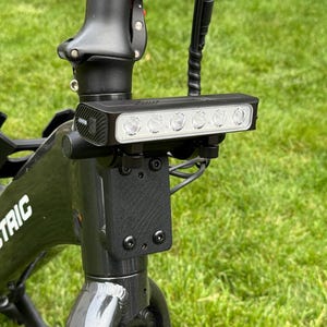 May include: Black electric bicycle with a bright LED headlight mounted on the handlebars. The headlight has five individual lights. The bicycle frame is black, and the background is green grass.