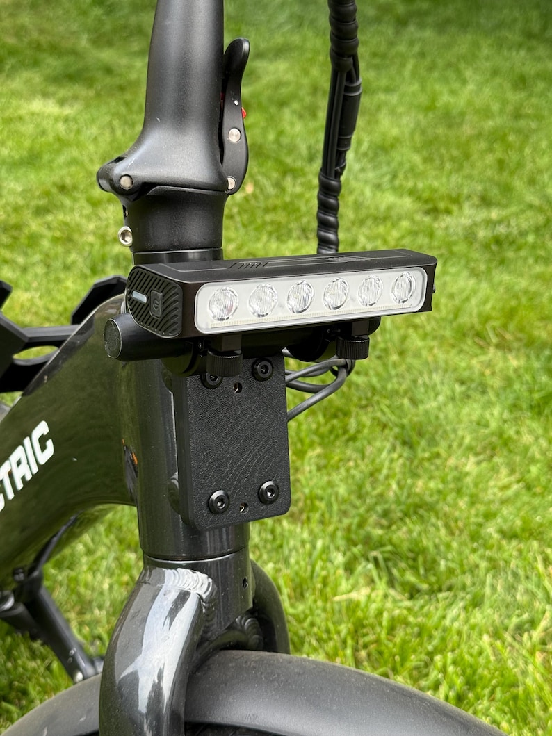 May include: Black electric bicycle with a bright LED headlight mounted on the handlebars. The headlight has five individual lights. The bicycle frame is black, and the background is green grass.