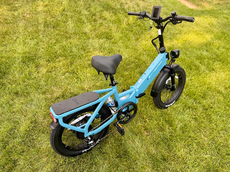 May include: A light blue Lectric XP 2.0 electric bike with black tires and a black seat is parked on a grassy lawn. The bike has a rear cargo rack, a digital display, and a headlight. The bike's frame is a vibrant blue.