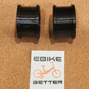 Op de afbeelding: Twee zwarte plastic ringen, waarschijnlijk voor een fiets, op een bruine kurk achtergrond. De ringen zijn glad en cilindrisch. Een sticker met de tekst "EBike BETTER" en een silhouet van een e-bike bevindt zich in het onderste midden van de afbeelding.