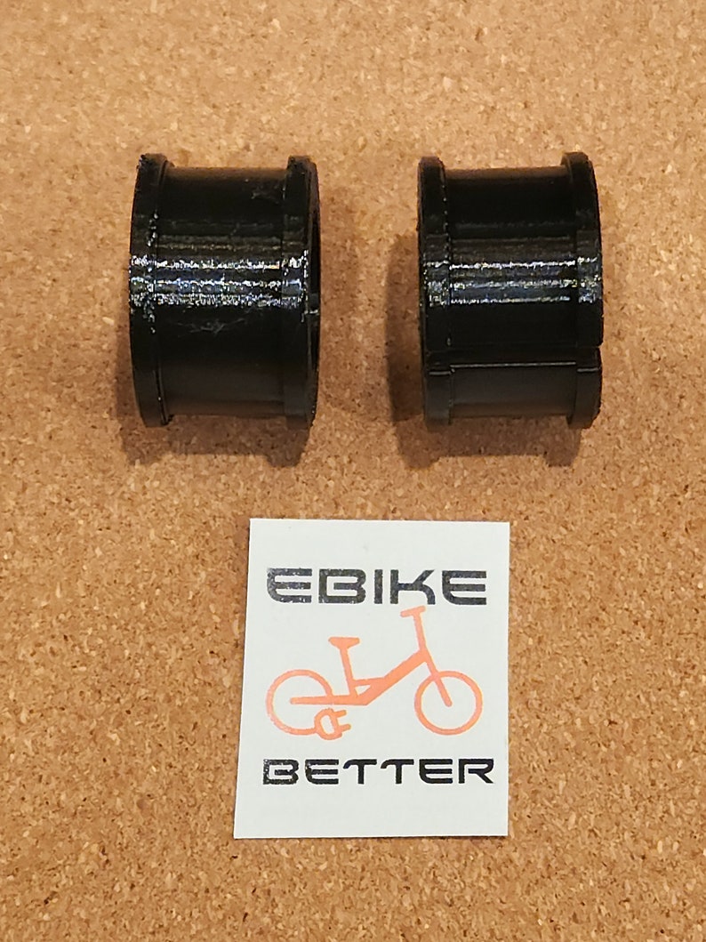 Op de afbeelding: Twee zwarte plastic ringen, waarschijnlijk voor een fiets, op een bruine kurk achtergrond. De ringen zijn glad en cilindrisch. Een sticker met de tekst "EBike BETTER" en een silhouet van een e-bike bevindt zich in het onderste midden van de afbeelding.