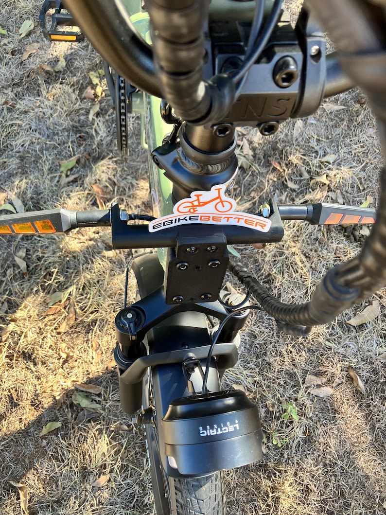 May include: An overhead view of an electric bicycle, showcasing the handlebars, front suspension, and headlight. The handlebars have orange reflectors and a sticker that says "eBikebetter". The headlight has the word "Lectric" printed on it.
