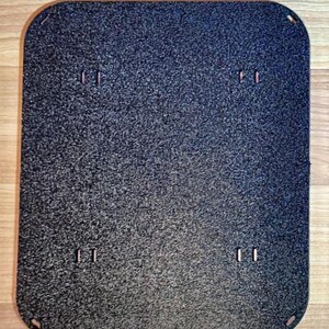 May include: A rectangular, dark gray textured plate with rounded corners. The plate has four pairs of small, rectangular cutouts and four small holes at the corners. The surface has a speckled appearance.