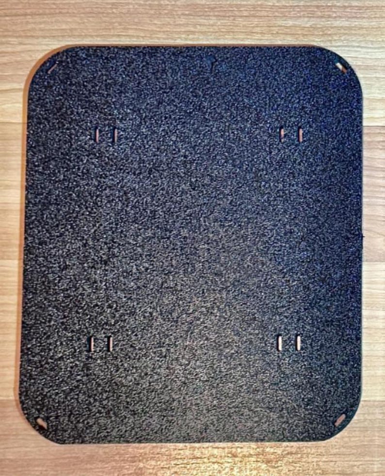 May include: A rectangular, dark gray textured plate with rounded corners. The plate has four pairs of small, rectangular cutouts and four small holes at the corners. The surface has a speckled appearance.