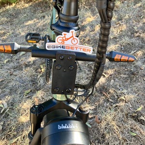 May include: Close-up of an electric bicycle with a black frame and a round headlight. The handlebars have orange turn signals and a sticker that says "EBIKEbetter". The bike is outdoors on a sunny day.