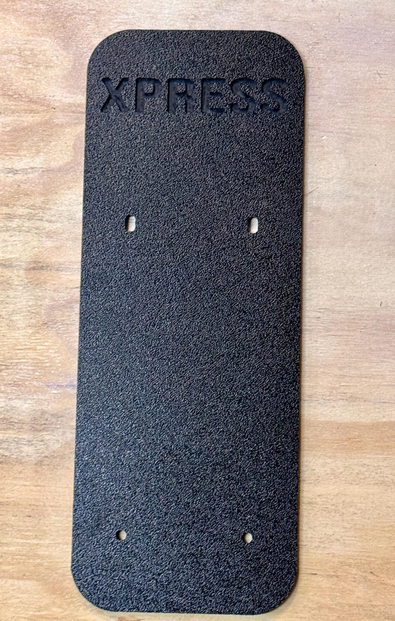 May include: Black rectangular plate with the word "XPRESS" embossed in the top centre. The plate has a textured surface and four small holes.