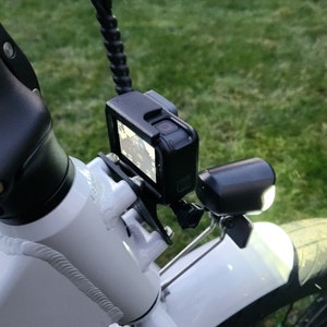 May include: A white bicycle with a black action camera mounted on the handlebars. The camera is in a protective case and has a red recording light on. There is a black light mounted on the front fender.