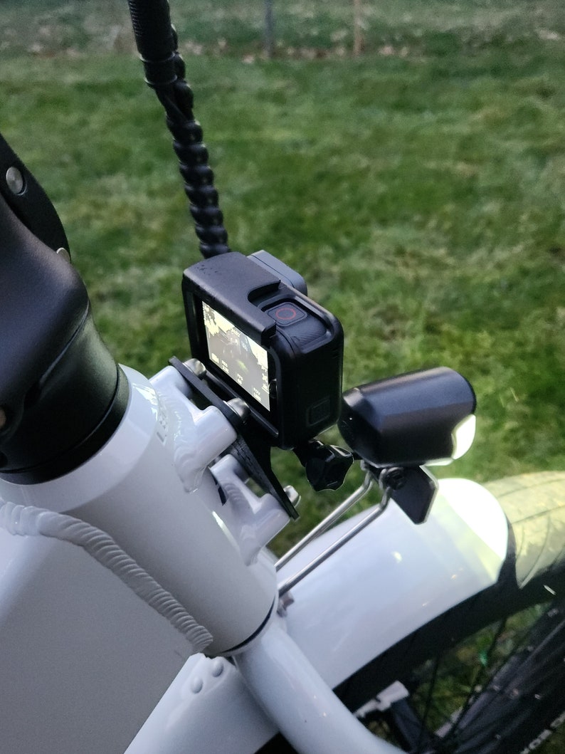 May include: A white bicycle with a black action camera mounted on the handlebars. The camera is in a protective case and has a red recording light on. There is a black light mounted on the front fender.