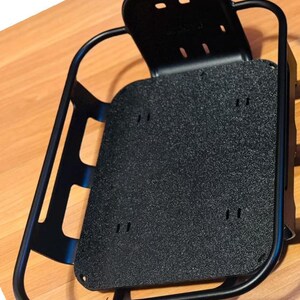 May include: A black metal bicycle rack with a textured, rectangular base and a curved frame. The rack has a back support with two holes and several mounting holes. The rack is designed to attach to a bicycle.