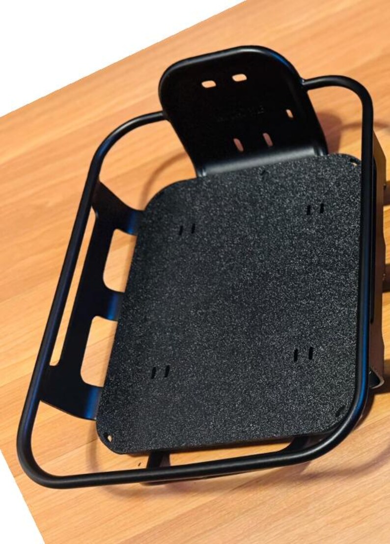 May include: A black metal bicycle rack with a textured, rectangular base and a curved frame. The rack has a back support with two holes and several mounting holes. The rack is designed to attach to a bicycle.