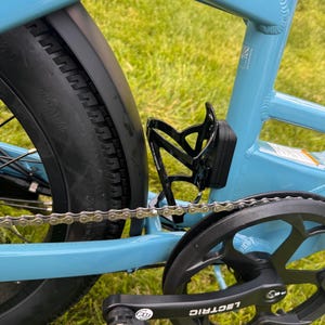 May include: Close-up of a light blue bicycle frame with a black tire, chain, and pedal. A black water bottle holder is attached to the frame. The word "LECTRIC" is visible on the pedal.
