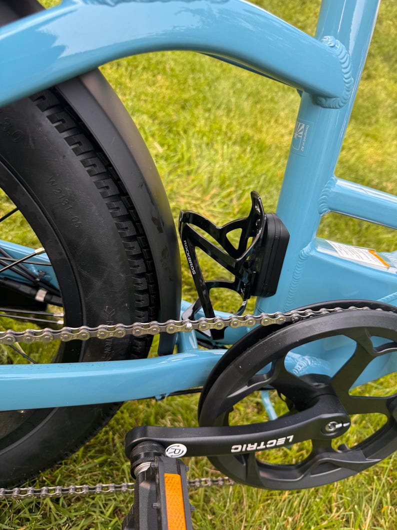 May include: Close-up of a light blue bicycle frame with a black tire, chain, and pedal. A black water bottle holder is attached to the frame. The word "LECTRIC" is visible on the pedal.
