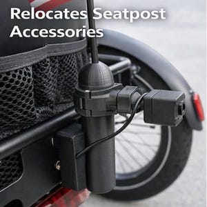 May include: Black bicycle accessory with the text "Relocates Seatpost Accessories." The accessory includes a cylindrical component, a rectangular box, and a camera-like device, all connected by wires. An American flag is visible in the background.