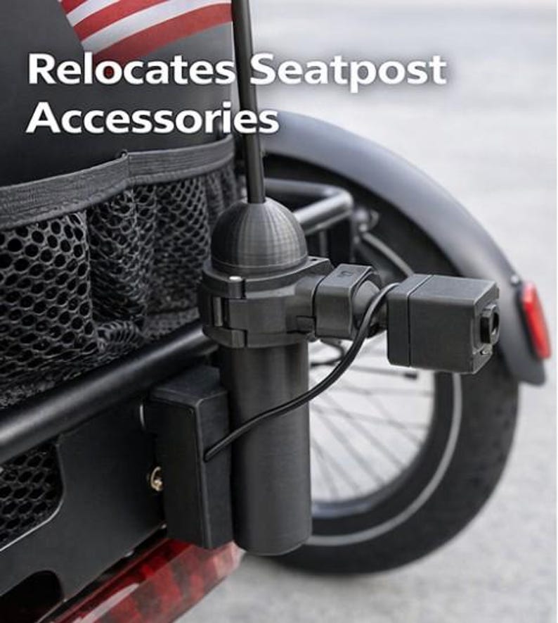 May include: Black bicycle accessory with the text "Relocates Seatpost Accessories." The accessory includes a cylindrical component, a rectangular box, and a camera-like device, all connected by wires. An American flag is visible in the background.