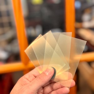 May include: A hand holding a stack of translucent, square-shaped plastic sheets. The sheets are slightly yellowed and appear to be thin. The background includes an orange chair and blurred elements.