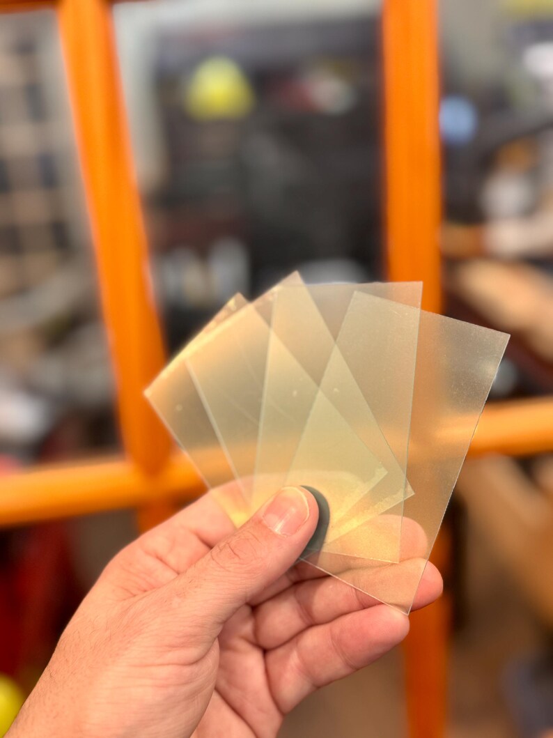 May include: A hand holding a stack of translucent, square-shaped plastic sheets. The sheets are slightly yellowed and appear to be thin. The background includes an orange chair and blurred elements.