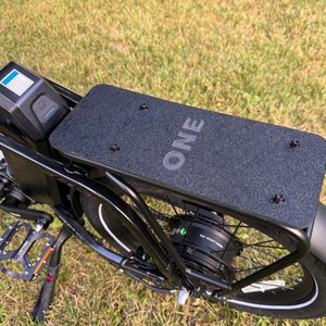 May include: Black electric bicycle with a black rear rack that says "ONE" on it. The bicycle has a black fender and a black tire with a white stripe.