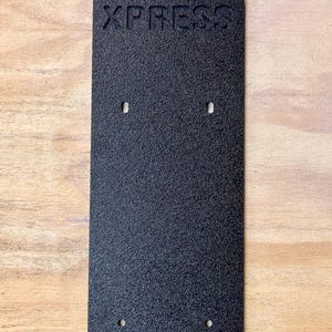 May include: Black rectangular plate with the word "XPRESS" embossed on the top. The plate has four holes drilled into it.