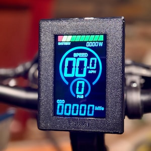 May include: A close-up of an electric bike display with a black frame. The screen shows a blue digital display with speed, battery life, and other metrics. The speed reads 00.0 MPH, and the odometer reads 00000 Mile.