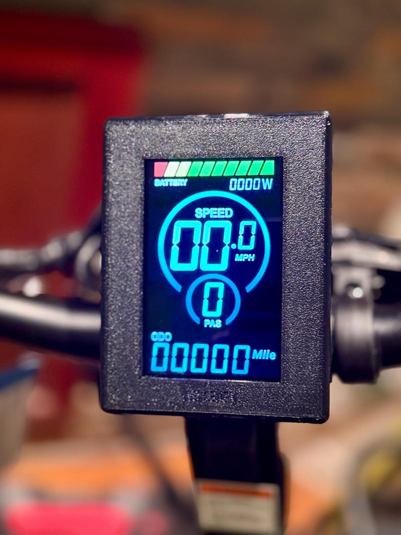 May include: A close-up of an electric bike display with a black frame. The screen shows a blue digital display with speed, battery life, and other metrics. The speed reads 00.0 MPH, and the odometer reads 00000 Mile.