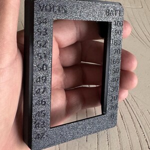 May include: A black rectangular frame with the words "VOLTS" and "BATT" printed on it. The frame has a textured surface and displays voltage and battery percentage markings from 45 to 54 and 10 to 100.