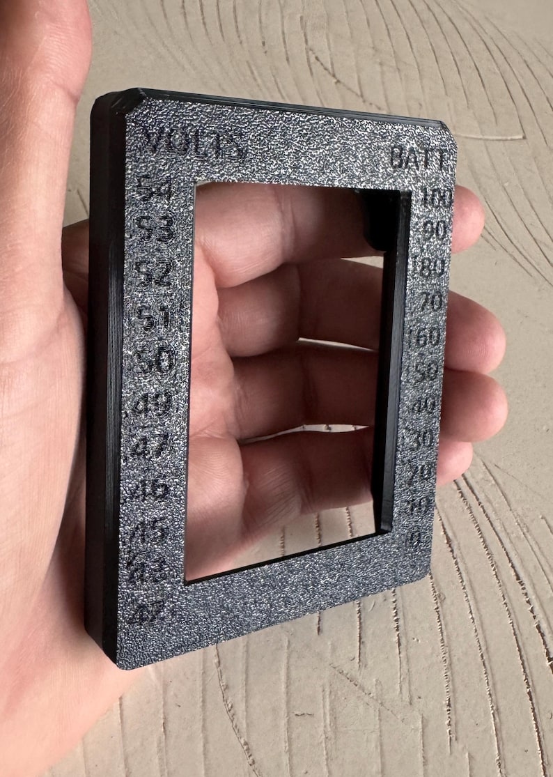 May include: A black rectangular frame with the words "VOLTS" and "BATT" printed on it. The frame has a textured surface and displays voltage and battery percentage markings from 45 to 54 and 10 to 100.