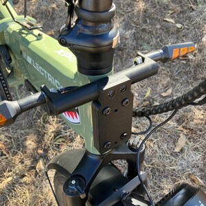 May include: Close-up of an olive green Lectric XP electric bike. The handlebars, stem, and front fork are black. The bike has a shark mouth graphic and orange turn signals. The tires are black.
