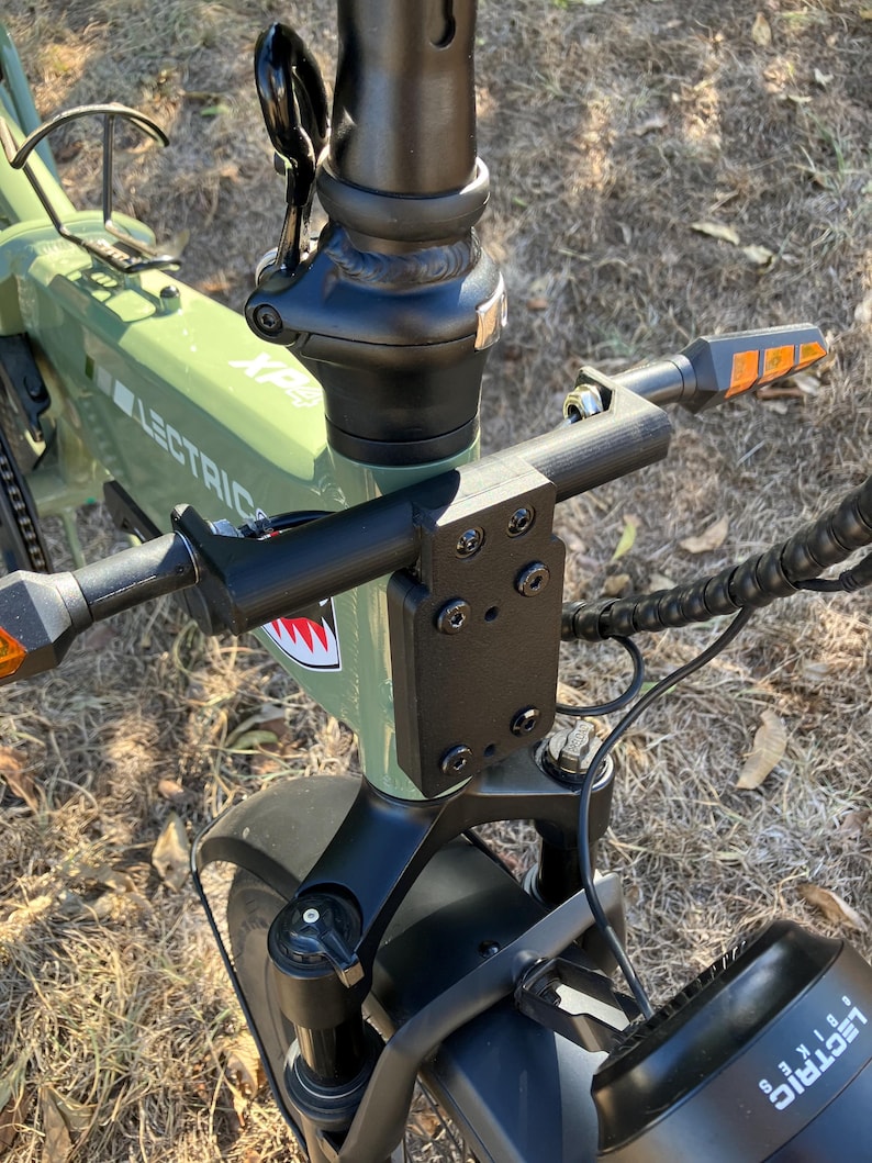 May include: Close-up of an olive green Lectric XP electric bike. The handlebars, stem, and front fork are black. The bike has a shark mouth graphic and orange turn signals. The tires are black.