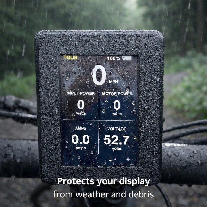 May include: A black digital display screen with data readings, including speed, power, and voltage, is shown. The screen is encased in a textured black frame, with the text "Protects your display from weather and debris" at the bottom. The display is wet with raindrops.