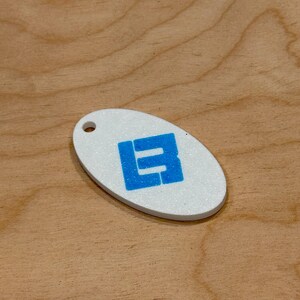 May include: White oval keychain with a blue block letter design. The keychain has a hole at the top for attachment. The background is a light brown wood grain.