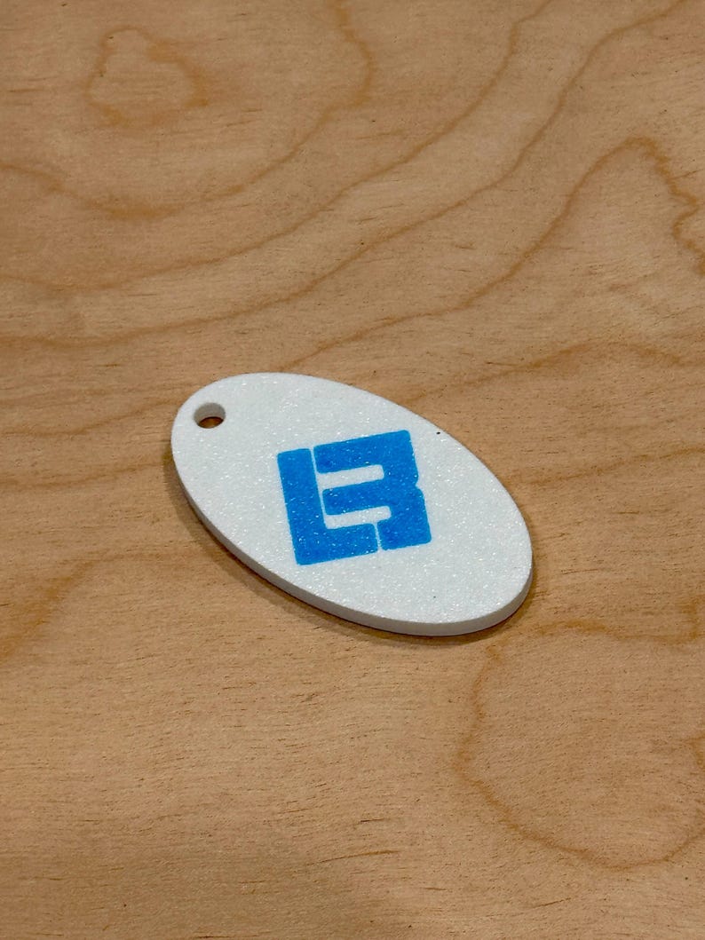 May include: White oval keychain with a blue block letter design. The keychain has a hole at the top for attachment. The background is a light brown wood grain.
