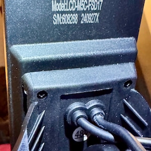 May include: Close-up of a black electronic component with the text "Model: LCD-M5C-FSD17" and "S/N:608268 240927X" printed in white. Two black cables are connected to the component. A sticker with the number "48" is visible.